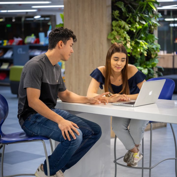 Dos personas sentadas en una mesa alta, interactuando frente a un computador portátil en un espacio interior moderno con plantas y estanterías al fondo.