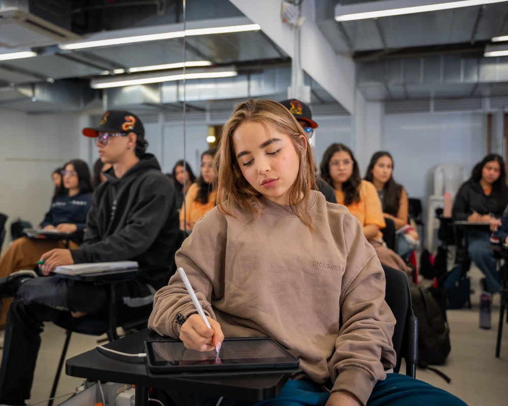 Varias personas sentadas en un aula, algunas tomando notas y otras usando dispositivos electrónicos sobre escritorios individuales.