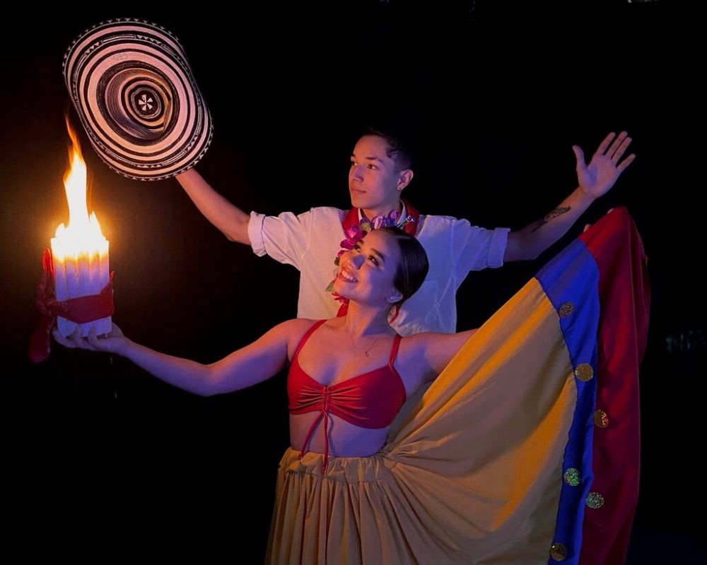 Dos personas en traje típico colombiano, una sostiene una vela encendida y la otra un sombrero vueltiao, con una bandera tricolor extendida.