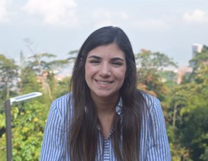 Persona con cabello largo y suelto viste una camisa de rayas azules y blancas, en un entorno exterior con vegetación y cielo parcialmente nublado al fondo.