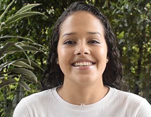 Persona con cabello oscuro y ondulado, vestida con camiseta blanca, está en un entorno exterior con vegetación en el fondo.