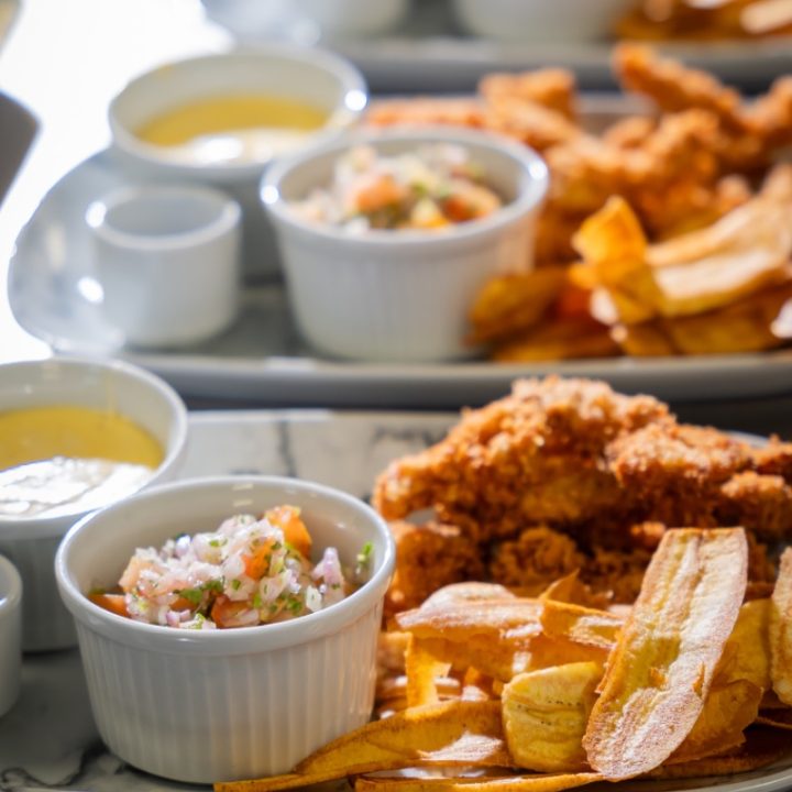 Plato servido con trozos de pollo empanizado, tajadas de plátano frito y una porción de ensalada en recipiente pequeño, acompañado de una salsa en taza blanca.