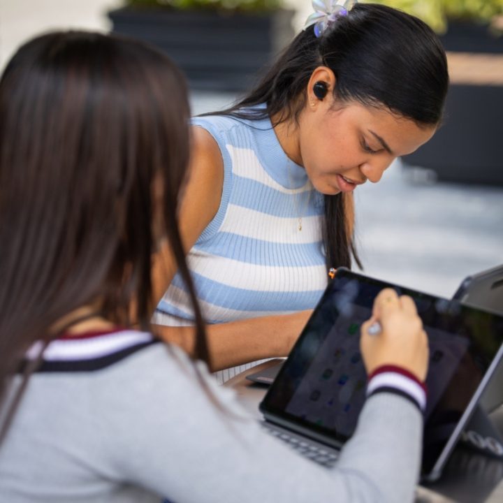 Dos personas trabajan en una mesa al aire libre; una utiliza un dispositivo tipo tableta con lápiz digital y la otra está frente a un computador portátil.