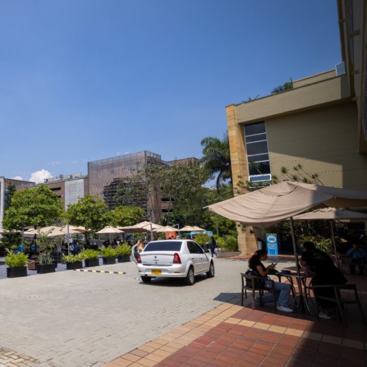 Zona exterior con mesas y sombrillas junto a un edificio, donde varias personas están sentadas; se observa un automóvil blanco estacionado y vegetación en el fondo bajo un cielo despejado.