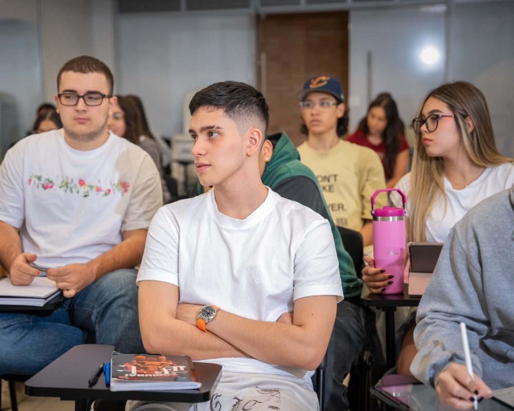 Varias personas sentadas en un aula, con escritorios individuales y cuadernos, participando en una clase.