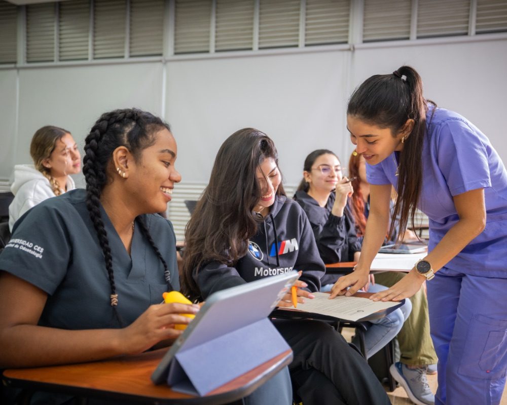 Varias personas en un aula interactúan durante una actividad; una persona de pie señala algo en un cuaderno mientras otras observan y toman notas, con tabletas y materiales sobre los escritorios.