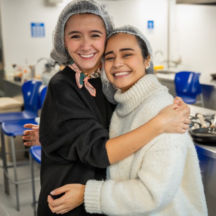 Dos personas con gorros de malla abrazadas en una cocina, con utensilios y sillas azules visibles en el fondo.