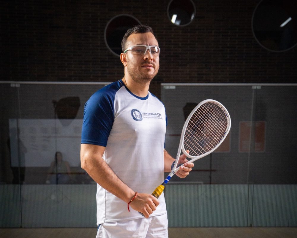 Persona en una cancha de squash sosteniendo una raqueta, vestida con camiseta deportiva blanca con mangas azules y pantalón corto blanco.