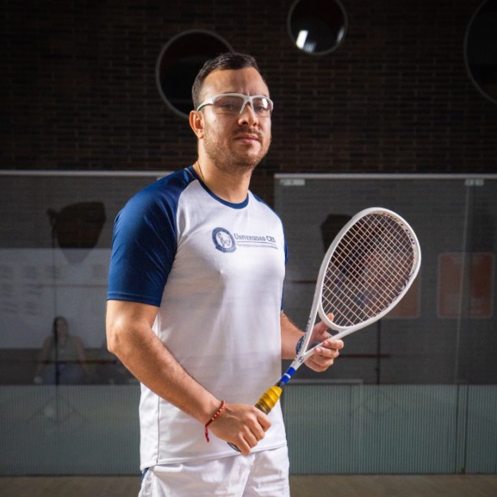 Persona en una cancha de squash sosteniendo una raqueta, vestida con camiseta deportiva blanca con mangas azules y pantalón corto blanco.