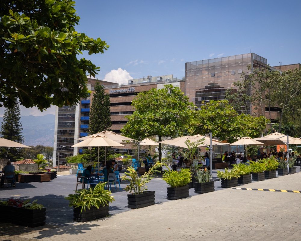 Zona al aire libre con mesas y sombrillas rodeadas de plantas, ubicada frente a edificios modernos con fachadas de vidrio y vegetación.