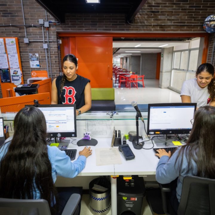 Área de atención con dos personas trabajando en computadores detrás de un mostrador, mientras otras dos personas están de pie frente a ellos. El espacio tiene paredes de ladrillo, una puerta naranja y mesas rojas visibles al fondo.