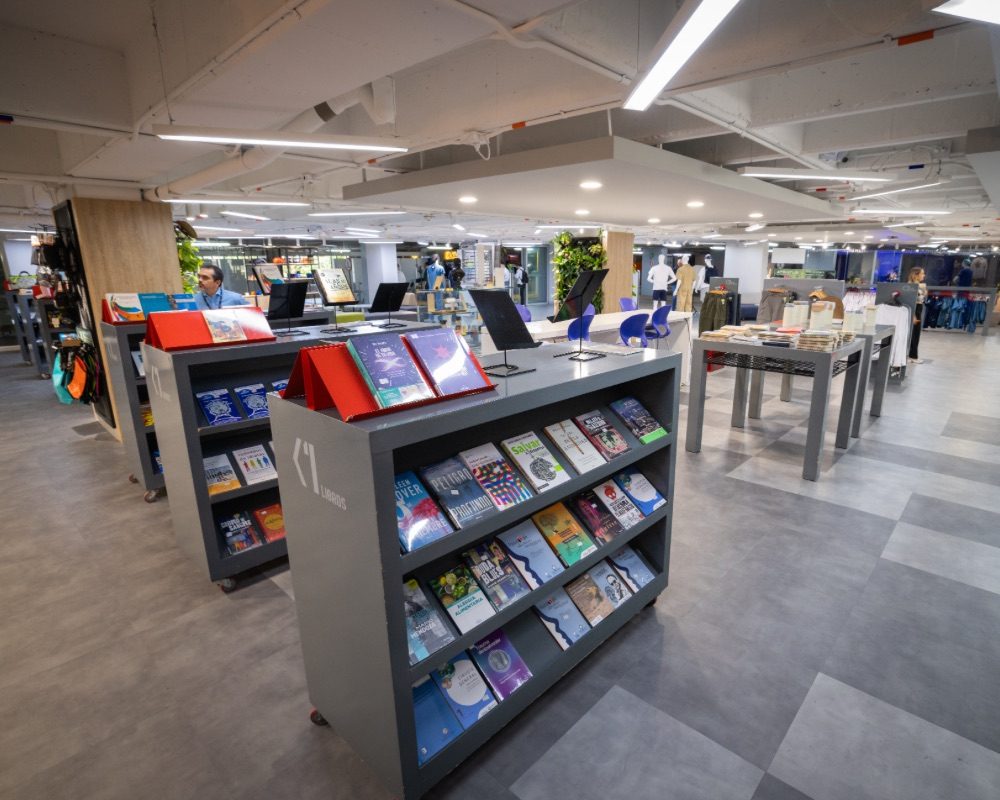 Espacio interior en tienda académica con estanterías que exhiben libros y materiales, mesas con productos organizados y una iluminación blanca uniforme.