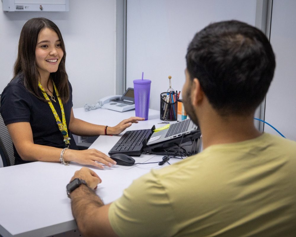 Dos personas sentadas frente a frente en un escritorio, con una computadora, teclado, teléfono y vaso sobre la mesa, en un espacio de oficina.