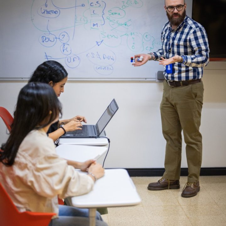 Persona de pie frente a un tablero con anotaciones, sosteniendo marcadores, mientras dos personas sentadas toman notas y usan un computador portátil.