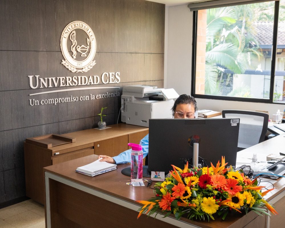Oficina con escritorio que tiene un computador, documentos y un arreglo floral colorido; en la pared se observa el logo y el nombre “Universidad CES” junto al lema institucional.