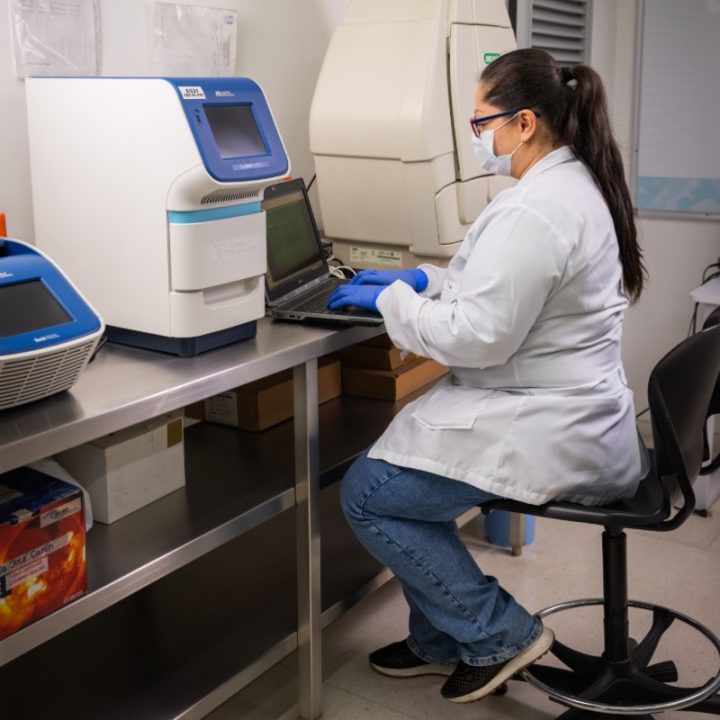 Persona con bata de laboratorio y guantes azules trabaja en una mesa utilizando un computador portátil, junto a equipos especializados en un entorno científico.