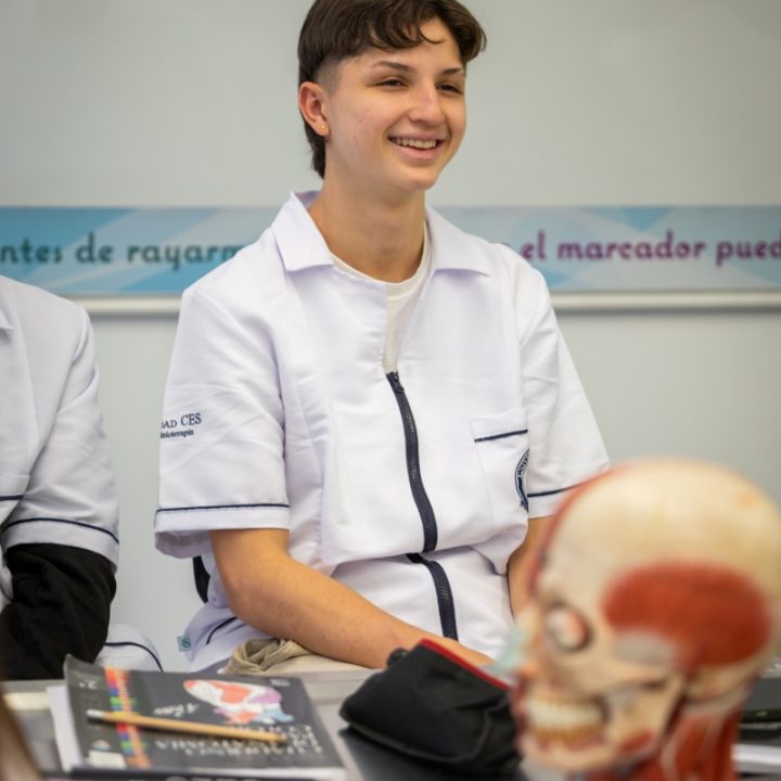 Persona con uniforme clínico está sentada en un aula frente a una mesa que tiene libros y un modelo anatómico de la cabeza humana.