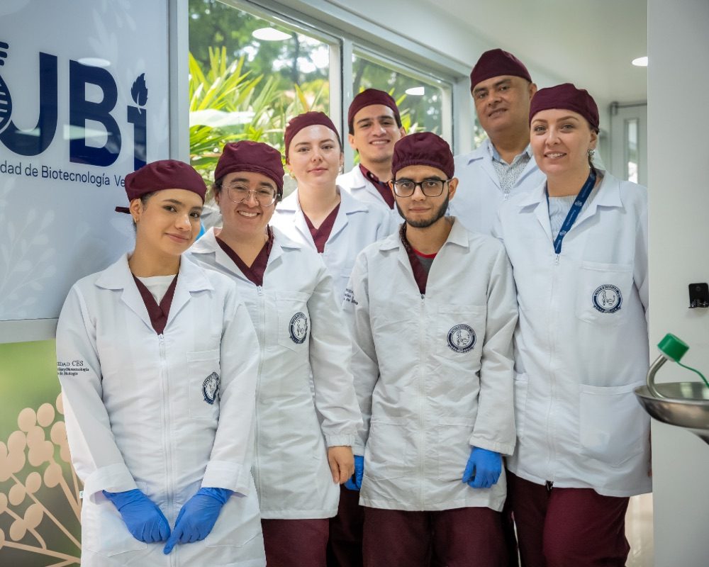 Grupo de personas con uniformes clínicos y guantes azules posan en un pasillo junto a un cartel que muestra el texto “UBi”.