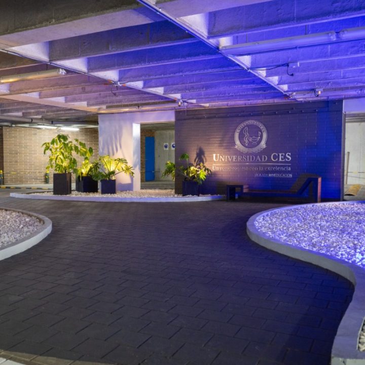 Entrada interior con piso oscuro y paredes claras, decorada con plantas y un gran panel que muestra el logotipo y nombre de la Universidad CES, iluminada con luz azul.