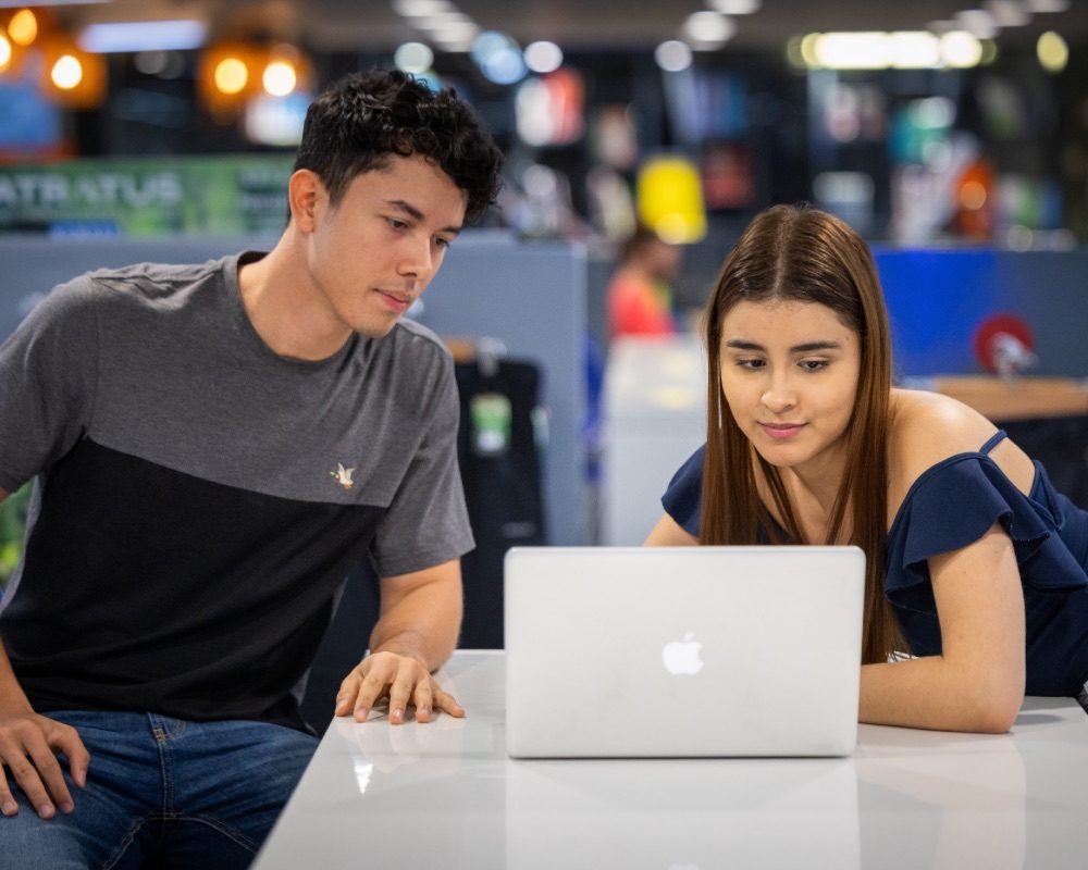 Dos personas en una mesa observan la pantalla de una computadora portátil en un espacio interior iluminado.