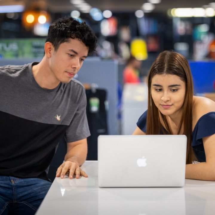 Dos personas en una mesa observan la pantalla de una computadora portátil en un espacio interior iluminado.