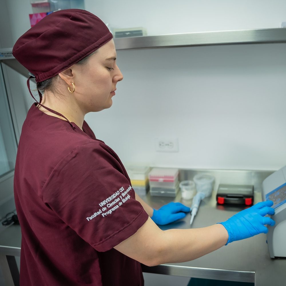 Persona con uniforme quirúrgico y guantes azules manipula material en una mesa de trabajo dentro de un laboratorio o área clínica, junto a equipos y recipientes utilizados para procedimientos médicos o de análisis.