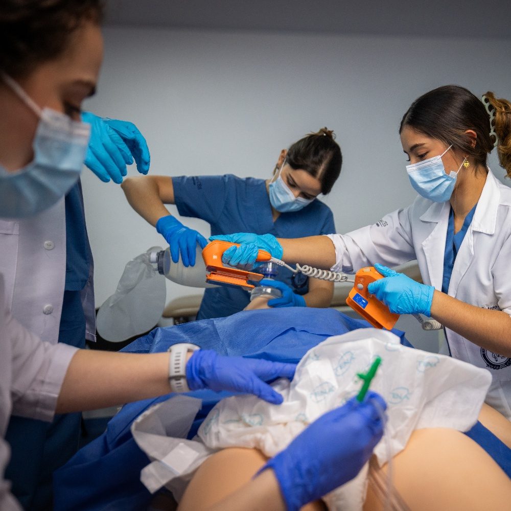 Varias personas con guantes y batas realizan una práctica médica sobre un simulador humano cubierto con una sábana azul; se observa el uso de un desfibrilador y otros instrumentos en un entorno clínico.