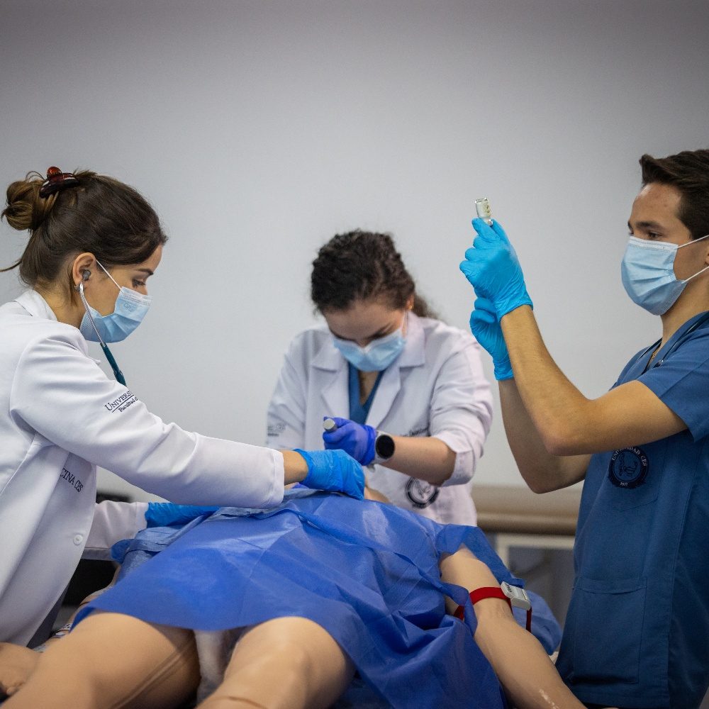 Varias personas con batas y guantes realizan un procedimiento médico simulado sobre un simulador humano cubierto con una sábana azul, utilizando instrumentos y jeringas en un entorno clínico.