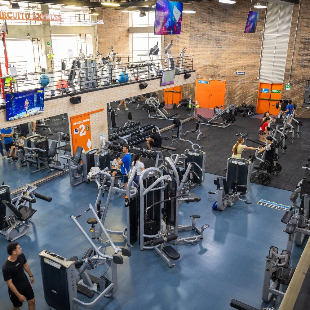 Vista interior de un gimnasio amplio con varias máquinas de ejercicio distribuidas en el espacio, algunas personas utilizando los equipos y una zona elevada con más implementos al fondo.