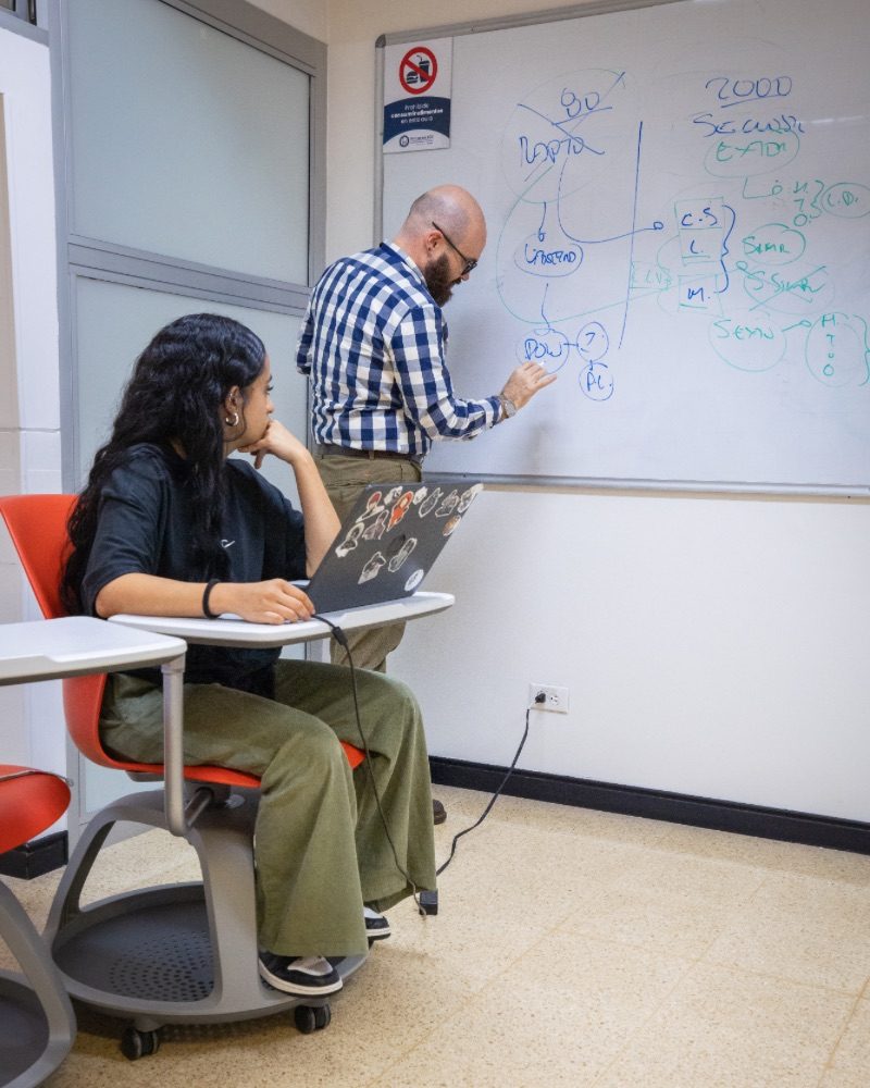 Una persona escribe en un tablero blanco diagramas y palabras mientras otra, sentada con un computador portátil, observa y toma notas en un aula o sala de clase.