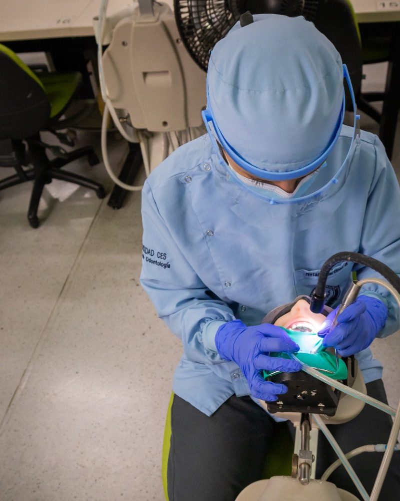 Persona con indumentaria odontológica y guantes azules realiza una práctica sobre un simulador dental utilizando instrumental en un entorno clínico equipado.
