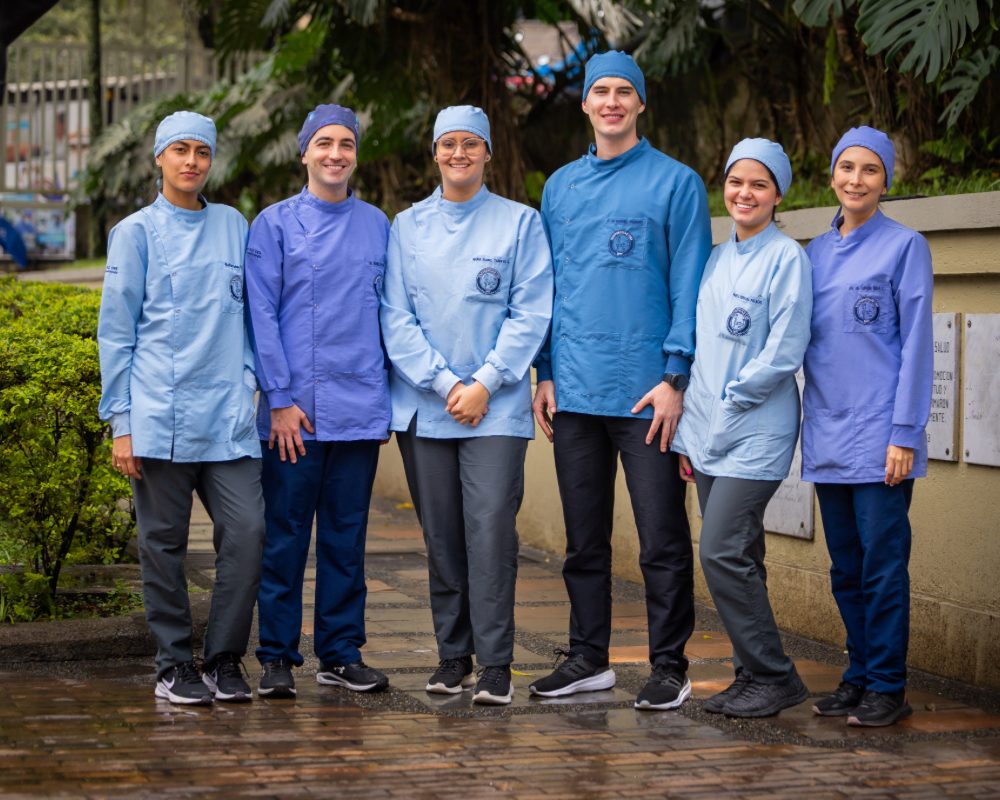 Seis personas de pie al aire libre, vestidas con uniformes clínicos en tonos azules y gorros quirúrgicos, posan juntas sobre un camino pavimentado con vegetación y árboles al fondo.