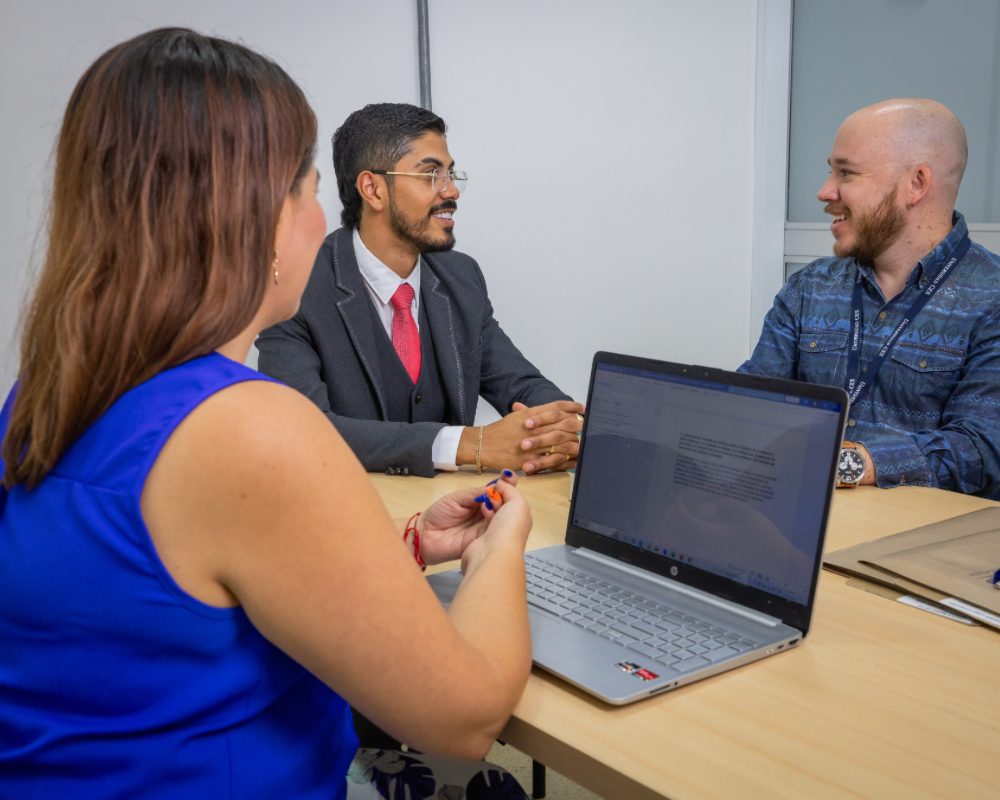 Tres personas sentadas alrededor de una mesa en una oficina, con un computador portátil abierto y una libreta sobre la superficie.