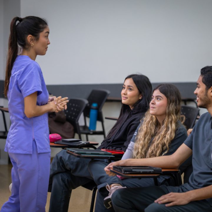Persona con uniforme clínico de color morado de pie frente a varias personas sentadas en un aula, con escritorios y sillas en el fondo.