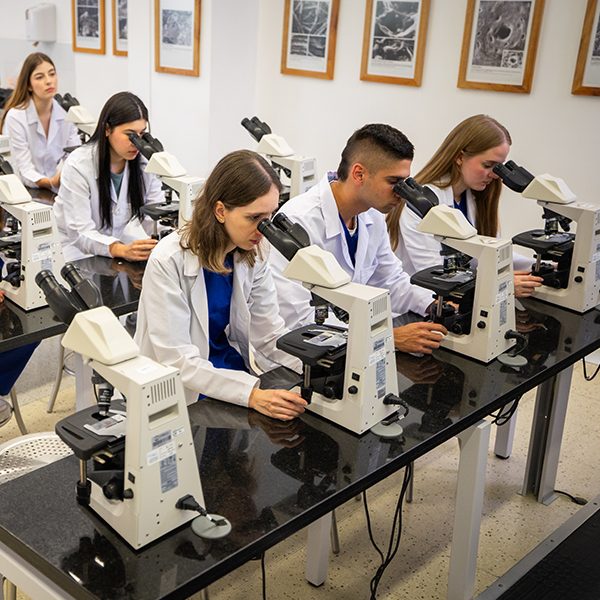 Varias personas con batas de laboratorio están sentadas en una mesa utilizando microscopios en un aula equipada para prácticas científicas. En la pared se observan cuadros con imágenes relacionadas con ciencia.