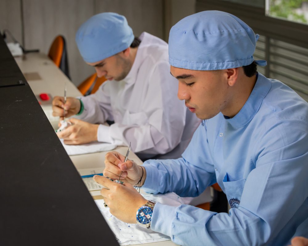 Dos personas trabajan en una mesa realizando prácticas manuales con instrumentos finos; ambas llevan gorros y batas de laboratorio en tonos claros.