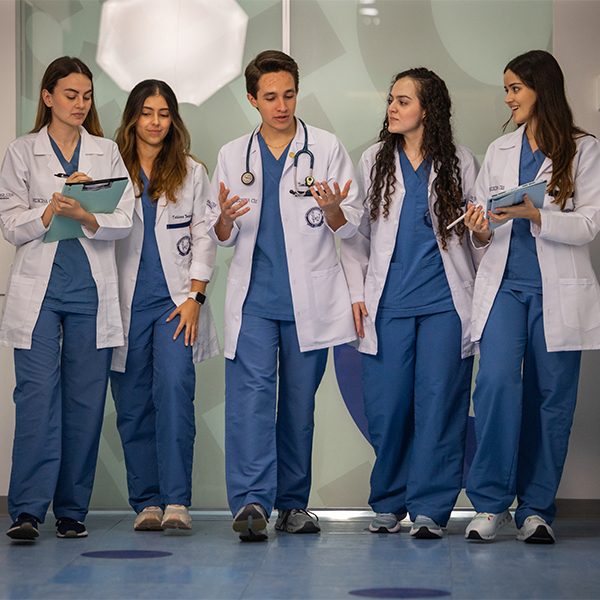 Cinco personas con uniformes médicos y batas blancas conversando y revisando notas en un pasillo clínico