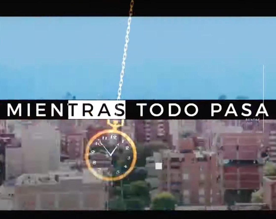 Imagen con fondo urbano y cielo azul. En el centro, sobre una franja negra horizontal, aparece el texto en mayúsculas: “MIENTRAS TODO PASA”. A la izquierda del texto se ve un reloj de bolsillo colgando con cadena dorada.