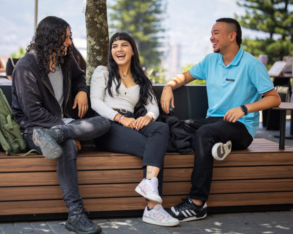 Tres personas están sentadas en una banca de madera al aire libre, conversando en un espacio con árboles y mesas en el fondo.