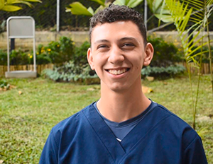 Persona con uniforme clínico azul está de pie en un espacio exterior con césped, plantas y una estructura metálica en el fondo.