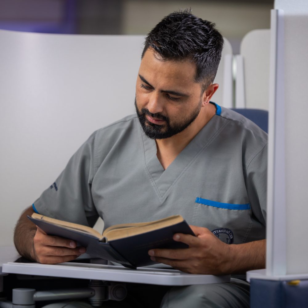 Persona con uniforme clínico está sentada leyendo un libro abierto en un espacio interior.