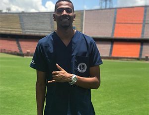 Persona con uniforme clínico azul está de pie sobre el césped de un estadio, con graderías multicolores visibles en el fondo.