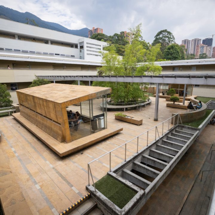 Terraza amplia con estructuras modernas de madera, áreas verdes y bancas, rodeada de edificios y con vista a montañas y ciudad al fondo.