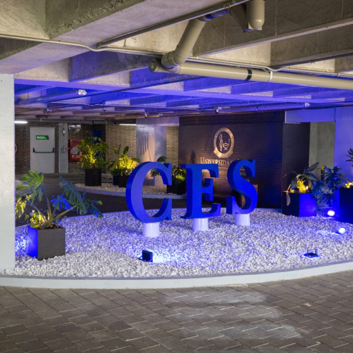 Zona interior con iluminación azul que resalta letras grandes formando “CES”, rodeadas de plantas decorativas y piedras blancas, junto a una pared con el logo institucional.