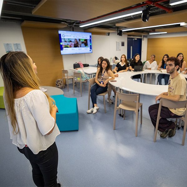 Una persona está de pie frente a un grupo en un aula moderna, dando una explicación. Varias personas están sentadas alrededor de una mesa circular, y en la pared hay una pantalla con contenido proyectado.