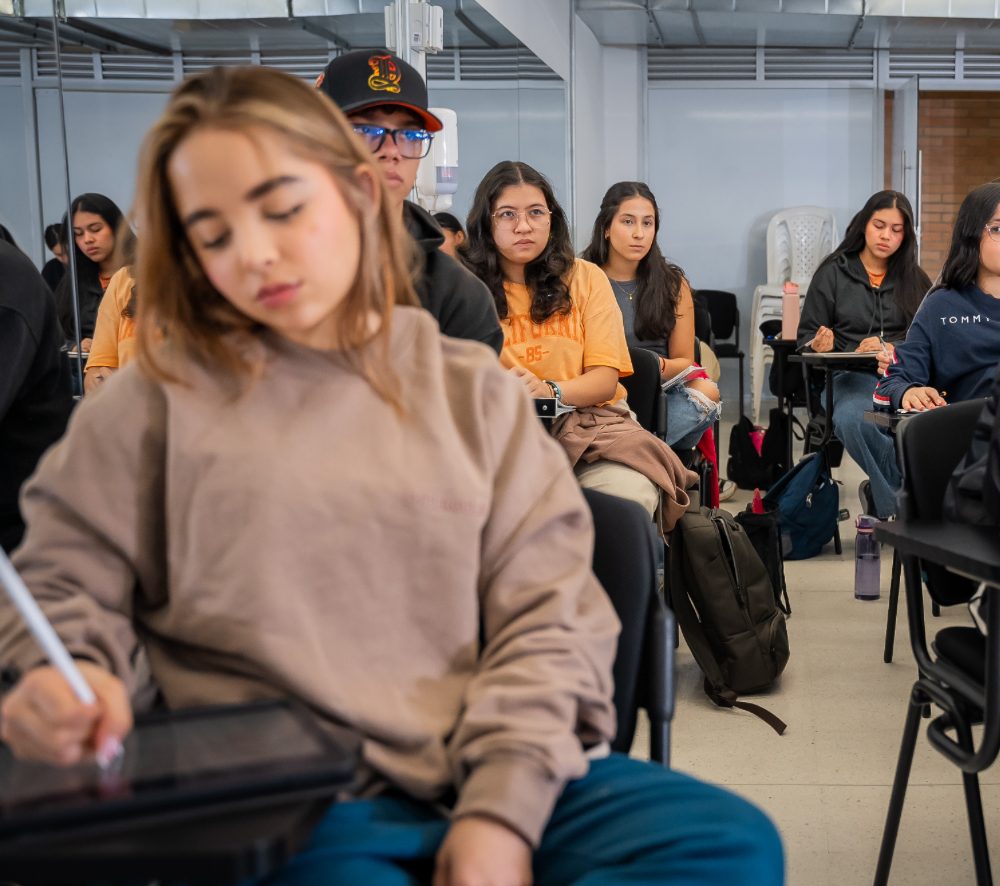 Varias personas están sentadas en un aula, tomando apuntes y usando dispositivos mientras atienden una clase; se observan escritorios, mochilas y un ambiente académico.
