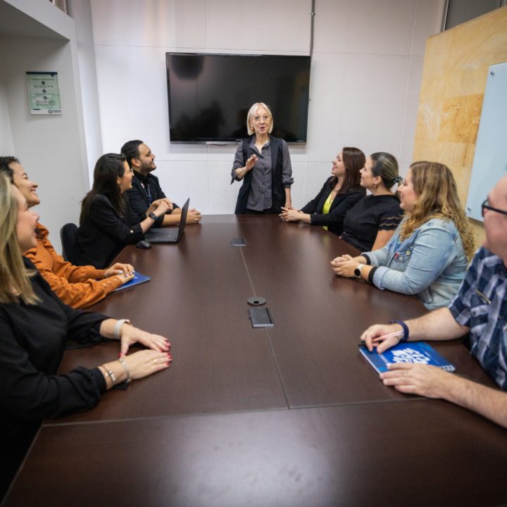 Varias personas sentadas alrededor de una mesa en una sala de reuniones, mientras otra persona está de pie hablando frente a una pantalla.