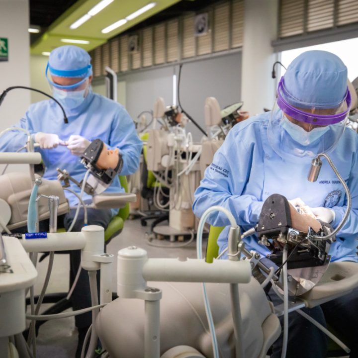 Personas con indumentaria quirúrgica realizan prácticas odontológicas en simuladores dentro de un laboratorio equipado con sillones y equipos dentales.