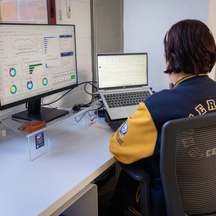 Persona sentada frente a un escritorio utilizando dos pantallas: un monitor con gráficos y tablas y una computadora portátil con texto.