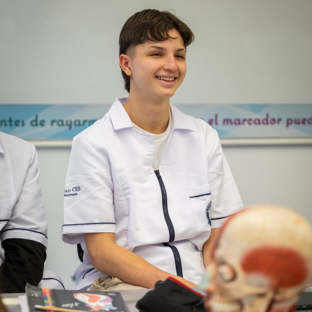 Persona con uniforme blanco sentado en un aula, sobre la mesa hay un modelo anatómico de cráneo y materiales de estudio.